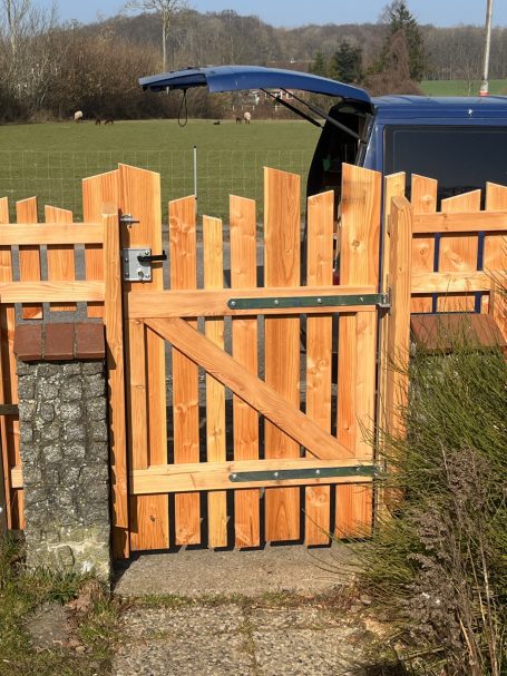 Holztor mit Zäunen und einem offenen Autofach auf einem sonnigen Feldweg.